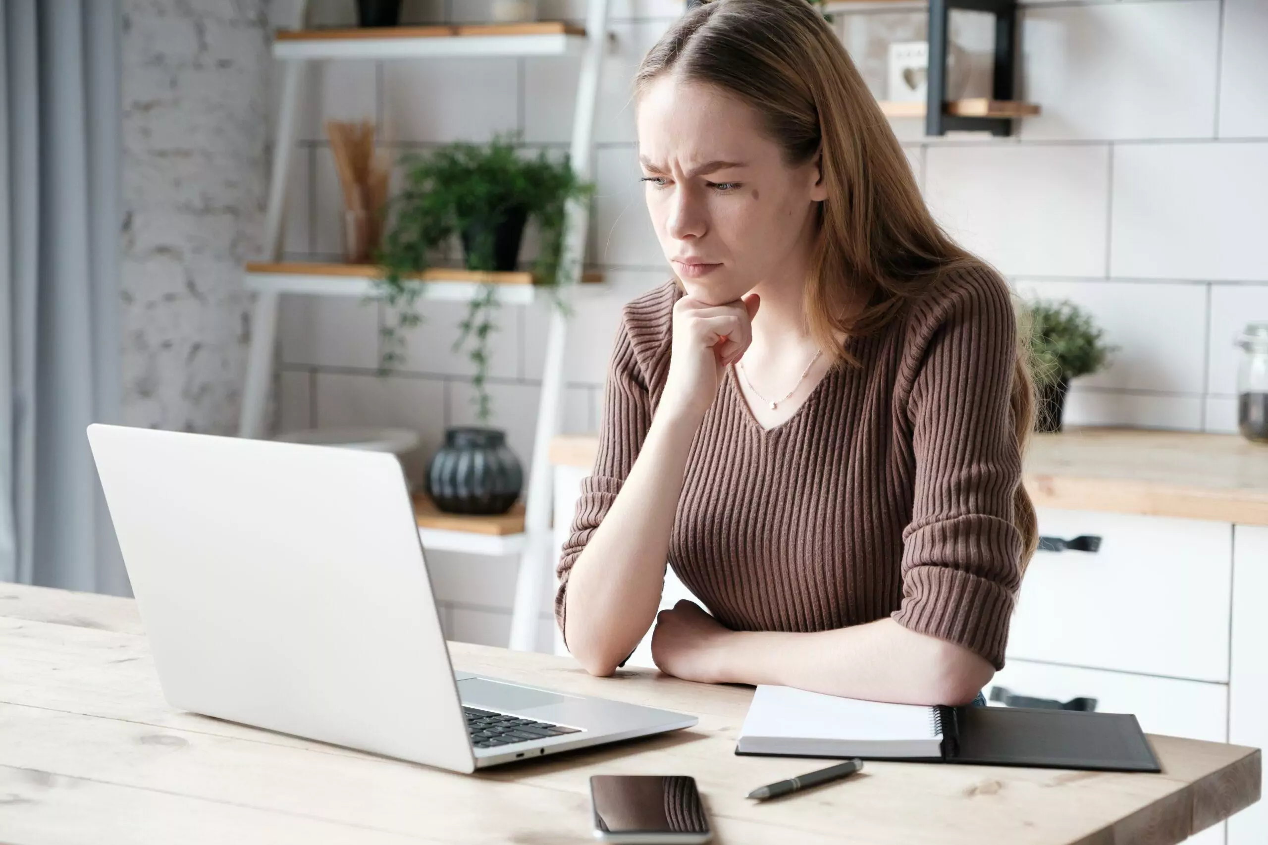Serious caucasian young female remote worker in casual outfit sitting at table with smartphone and planner and looking pensively at laptop screen