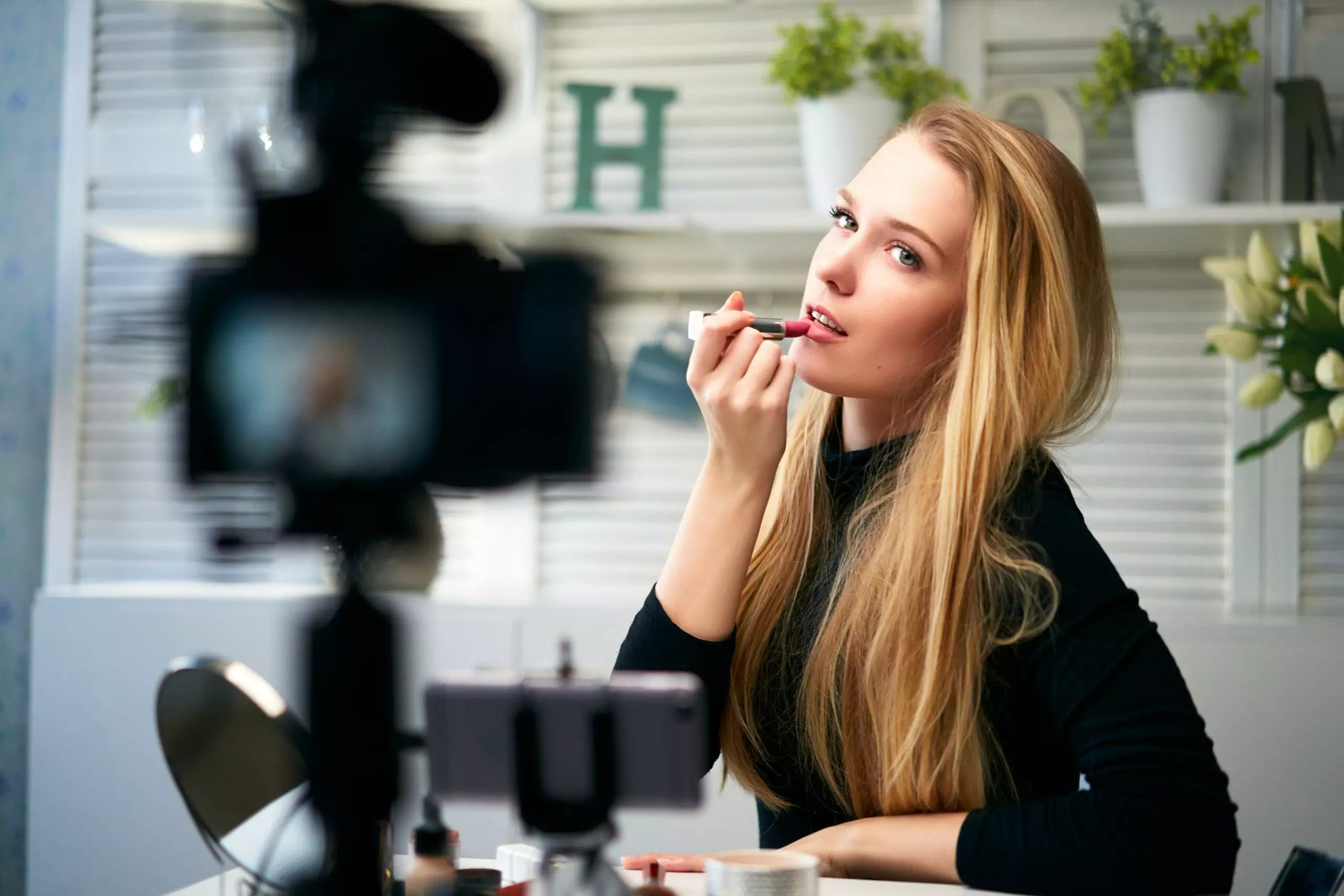 Vlogger female applies lipstick on lips. Beauty blogger woman filming daily make-up routine tutorial at camera on tripod. Influencer blonde girl live streaming cosmetics product review in home studio